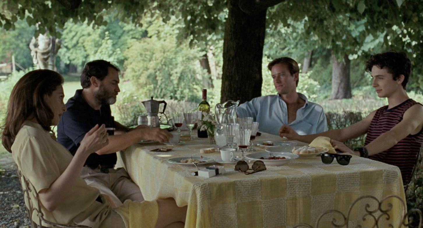 The Perlman family and Oliver eat lunch at a table under the trees, a classical statue in the garden behind them