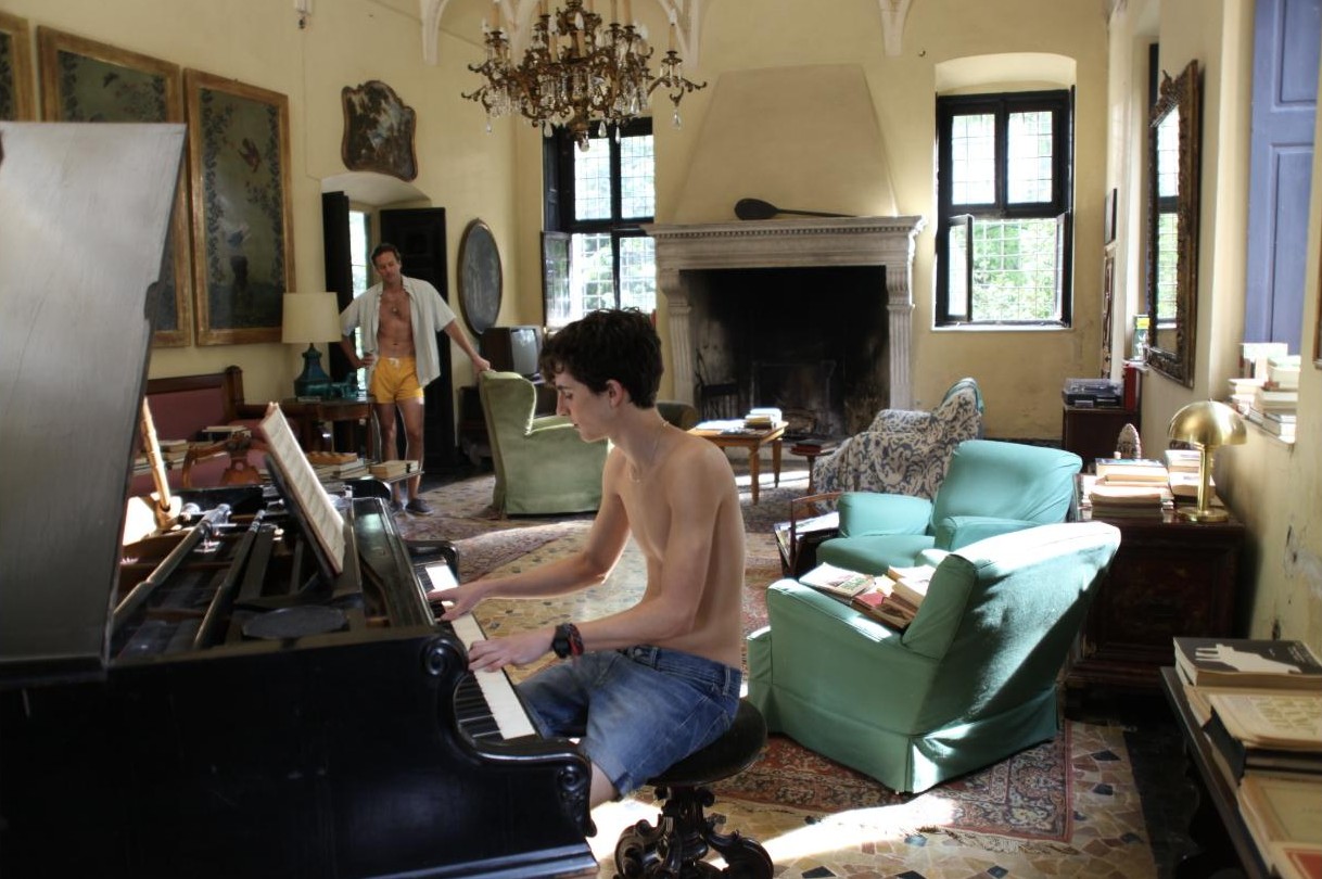 Elio plays the piano in the villa's living room while Oliver watches from the doorway